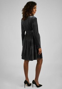 Abito in velluto scuro con maniche lunghe, cintura adatta in vita e gonna a ruota. Presenta righe verticali e una leggera lucentezza. Scarpe con tacco alto nere.