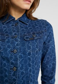 Denim jacket in dark blue with a textured geometric pattern, featuring a collar, button closure, and two front pockets with flap.