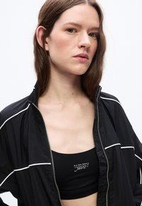 Jeune femme aux cheveux bruns portant une veste noire zippée par-dessus un haut court noir avec le logo "tommy jeans" sur un fond blanc uni.