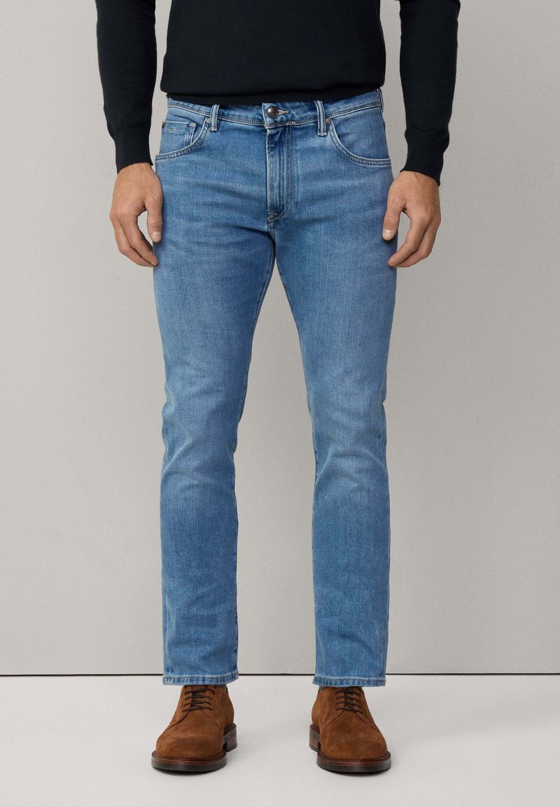 Jeans de denim azul claro con un corte recto, diseño de cinco bolsillos y cierre de botón de metal, combinados con zapatos de ante marrón.