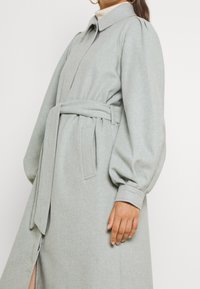 Manteau en laine gris clair avec collier, longues manches bouffantes, taille ceinturée et poches latérales. Texture lisse avec une coupe droite.