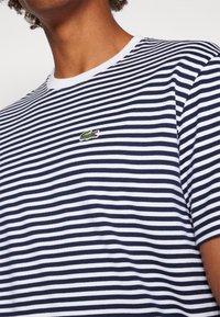 Person wearing a navy and white striped short-sleeve shirt with a small green crocodile logo on the chest.