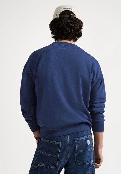 Μπλε ναυτικό φούτερ με ριμπ μανσέτες και τελείωμα, συνδυασμένο με σκουρόχρωμα τζιν, με μεγάλες πίσω τσέπες σε στιλ χρηστικότητας.