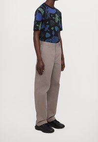 Mężczyzna w beżowych spodniacach o prostym kroju, czarnych sneakersach i czarnej koszulce z niebieskimi i zielonymi wzorami kwiatowymi, stojący na jednolitym tle.