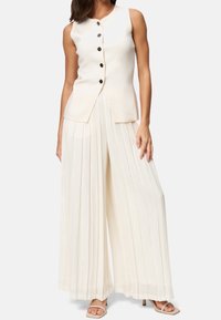 Femme portant un top sans manches crème à boutons associé à un pantalon large à taille haute plissé crème et des sandales à talons beiges.