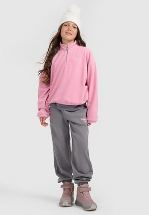 Meisje met een witte gebreide muts, een roze fleece trui, een grijze joggingbroek en roze winterlaarzen, staand tegen een effen achtergrond.