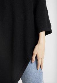 Main reposant sur un jean en denim bleu clair, portant un vêtement noir à manches longues ample avec un tissu texturé et un large poignet.