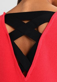 Czerwony dzianinowy top z głębokim dekoltem w kształcie V z krzyżującymi się czarnymi paskami, podkreślający teksturę i kontrastujące kolory. Miękki materiał o dopasowanym kroju.