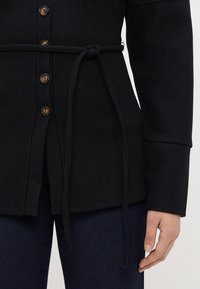 Svart beltejakke med knappelukking foran, lange ermer og glatt tekstur. Beltet er knyttet i midjen.