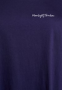 Mörkblå t-shirt i mjuk bomullstyg, med vit kursiv text "Moonlight Shadow" på övre vänstra sidan.