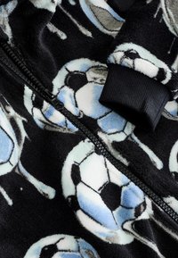 Svart fleecejacka med ett mönster av fotbollar i blått och vitt. Inkluderar en svart dragkedja och ribbade cuffs för en tät passform.