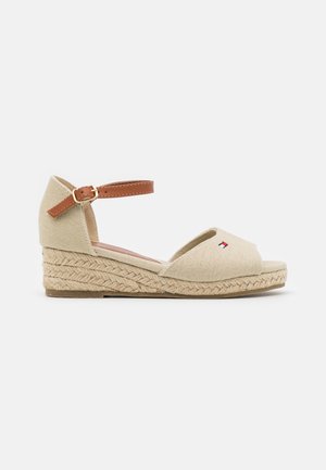 Beige kile-sandal med flettet rebssål, åben tå og brun ankelrem med guldspænde. Lille broderet logo på tå-remmen.