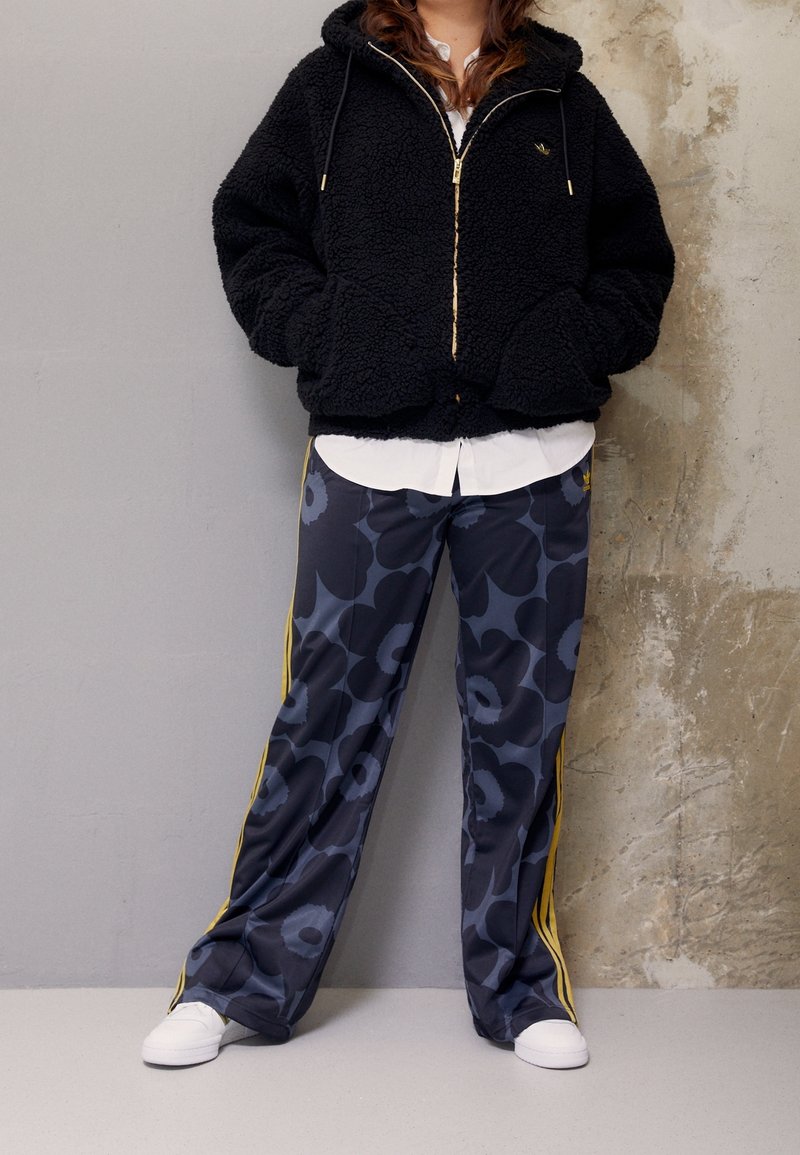 Sudadera con cremallera de forro polar negra con herrajes dorados, combinada con pantalones de chándal florales en navy con franjas amarillas a los lados y zapatillas blancas.