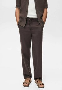 Dunkelbraune, leichte, weit geschnittene Hosen mit elastischem Bund und Kordelzug, kombiniert mit einem neutralen Shirt und braunen Sandalen.