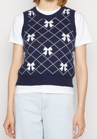 Marineblaue Strickweste mit weißen Schleifenmustern und Rautendesign. Rippstrickkragen und Saum. Über einem weißen T-Shirt getragen. Helle blaue Jeans sichtbar.