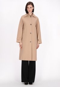 Beige Trenchcoat mit Kragen, Knopfleiste und langen Ärmeln. Verfügt über Taschen an den Seiten und wird mit schwarzen weiten Hosen kombiniert.