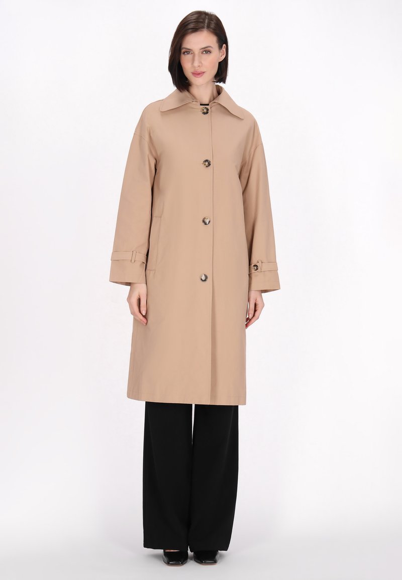 Beige Trenchcoat mit Kragen, Knopfleiste und langen Ärmeln. Verfügt über Taschen an den Seiten und wird mit schwarzen weiten Hosen kombiniert.