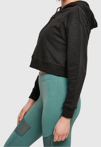 Felpa nera corta con cappuccio e cordino, abbinata a leggings teal con pannelli a rete e pannelli solidi. Tessuto morbido, design aderente e polsini a coste.