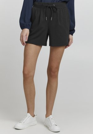 Pantaloni corti neri con vita elasticizzata e cordoncino, dotati di tasche laterali e una consistenza del tessuto morbida e liscia. Indossati con sneakers bianche.