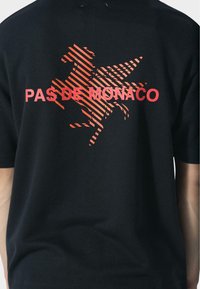 Zwarte t-shirt met rood diagonaal gestreepte paardenafbeelding en rode tekst "PAS DE MONACO" over het paard op de achterkant.