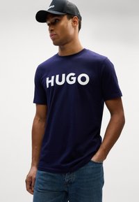 Tumši zilas krāsas kokvilnas t-krekls ar lielu baltu uzrakstu "HUGO" priekšā, apvienots ar melnu sietu cepuri. Modelis valkā zilos džinsus.