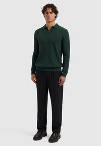 Groene gebreide pullover met een kraag en rits, gecombineerd met een zwarte broek en zwarte schoenen, staande tegen een neutrale achtergrond.