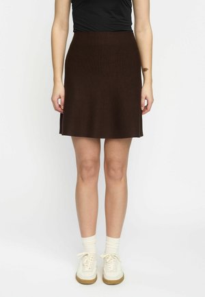 Brauner Strickrock mit weitem Schnitt und strukturierter Oberfläche, der über dem Knie endet. Kombiniert mit weißen Schuhen und cremefarbenen Socken. Schlichtes Design, keine sichtbaren Details.