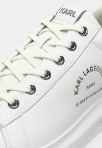 Hvit sneakers med kremfargede snører, metalløyler og "KARL LAGERFELD PARIS 21 RUE ST-GUILLAUME" trykt på siden.