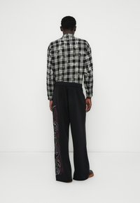 Πουκάμισο με ασπρόμαυρο καρό, μακριά μανίκια και κυρτή γωνία κάτω, συνδυασμένο με μαύρο παντελόνι που έχει κόκκινη και λευκή αφηρημένη κεντητική στη πλευρά.