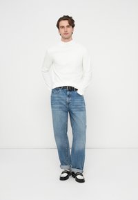 Biała koszula z długim rękawem i wysokim kołnierzem, w połączeniu z jasnoniebieskimi dżinsami i czarnym paskiem. Obuwie: czarne buty z białymi akcentami.