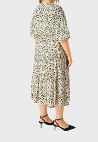 Šaty s květinovým vzorem v béžové a černé barvě, s nabíranými rukávy a skládací sukní, doplněné černými sandály na vysokém podpatku.