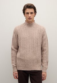 Maglione beige a treccia con collo alto, polsini e orlo a coste. La texture è morbida con motivi in rilievo su tutta la superficie. Indossato con pantaloni marroni scuri.