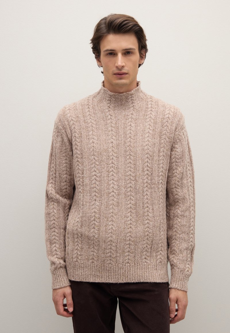 Maglione beige a treccia con collo alto, polsini e orlo a coste. La texture è morbida con motivi in rilievo su tutta la superficie. Indossato con pantaloni marroni scuri.