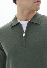 Zeleni pulover z teksturo in zadrgo na ovratniku, sicer pa s belim notranjim slojem, ki ima srebrno zadrgo. Pogled od blizu na ovratnik.