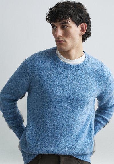 Blå, strikket sweater med runde udskæring og ribbede manchetter, med en struktureret overflade. Båret over en hvid skjorte, sat mod en lys baggrund.