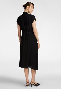 Vestito nero con bottoni, maniche corte arrotolate, vita svasata e gonna a-line fluente che termina a metà polpaccio, abbinato a tacchi neri.