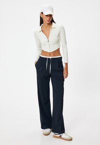 Weißes, geripptes Croptop mit Kragen und Reißverschluss, kombiniert mit lockeren, marineblauen Hosen mit weißen Kordeln und Taschen, abgerundet mit weißen Sneakers.