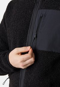Svart fleecejacka med en texturerad yta, som har en slät svart panel och dragkedja på bröstet, och en hand som håller i dragkedjans flik.