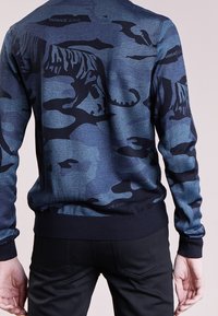 Pull camouflage bleu marine avec des silhouettes de tigres noirs, finitions côtelées au niveau des poignets et de l'ourlet, avec le nom de la marque "VERSACE JEANS" subtilement intégré.