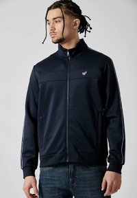 Veste zippée marine avec col montant, poches latérales et rayures blanches contrastantes sur les manches. Présente un logo sur la poitrine. Texture de tissu lisse.