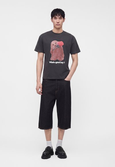 Jonge man die een zwart T-shirt draagt met een hond die een rode roos vasthoudt en de tekst "Who's a good boy?", zwarte knielange shorts, witte sokken en zwarte schoenen.