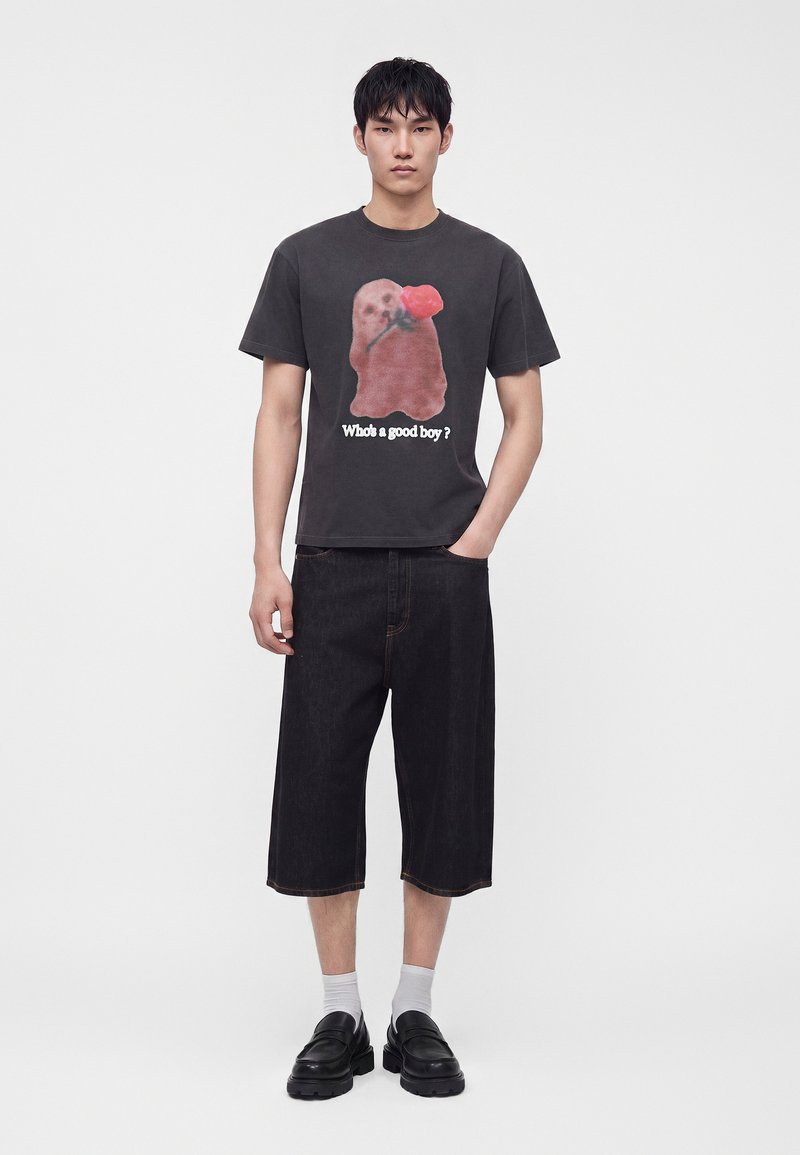 Νέος άνδρας που φοράει μαύρο T-shirt με έναν σκύλο που κρατάει μια κόκκινη τριανταφυλλιά και το κείμενο "Who's a good boy?", μαύρα σορτς μέχρι το γόνατο, λευκές κάλτσες και μαύρα παπούτσια.