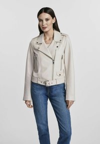 Jasnobeżowa skórzana kurtka motocyklowa z zamkami, paskiem w talii i ząbkowanym kołnierzem. Nosi się ją na niebieskich dżinsach, z długimi rękawami.