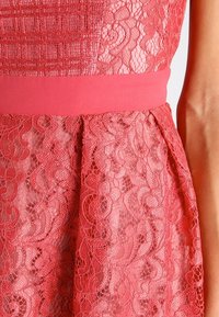 Röd spetsklänning med en texturerad överdel och ett slätt rosa midjeband. Kjolen har intrikata spetsmönster och en mjuk, genomskinlig textur.