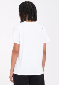 Camiseta de algodón blanca de mangas cortas, con cuello redondo y un pequeño detalle del logo en la parte de atrás, combinada con pantalones oscuros. Textura suave y corte relajado.