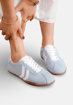 Personne portant un pantalon blanc et des baskets bleu clair pointe sa cheville avec ses deux mains ornées de bagues et de bracelets.