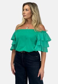 Zielony top bez ramiączek z warstwowymi, falbaniastymi rękawami. Gładki materiał, luźny krój, zestawiony z ciemnymi, wysokimi dżinsami. Prosta naszyjnik jako akcent.