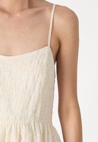 Vestido claro de crema con tirantes finos, que presenta un bordado floral texturizado y un dobladillo fruncido. Tejido suave y ligero con un patrón delicado.