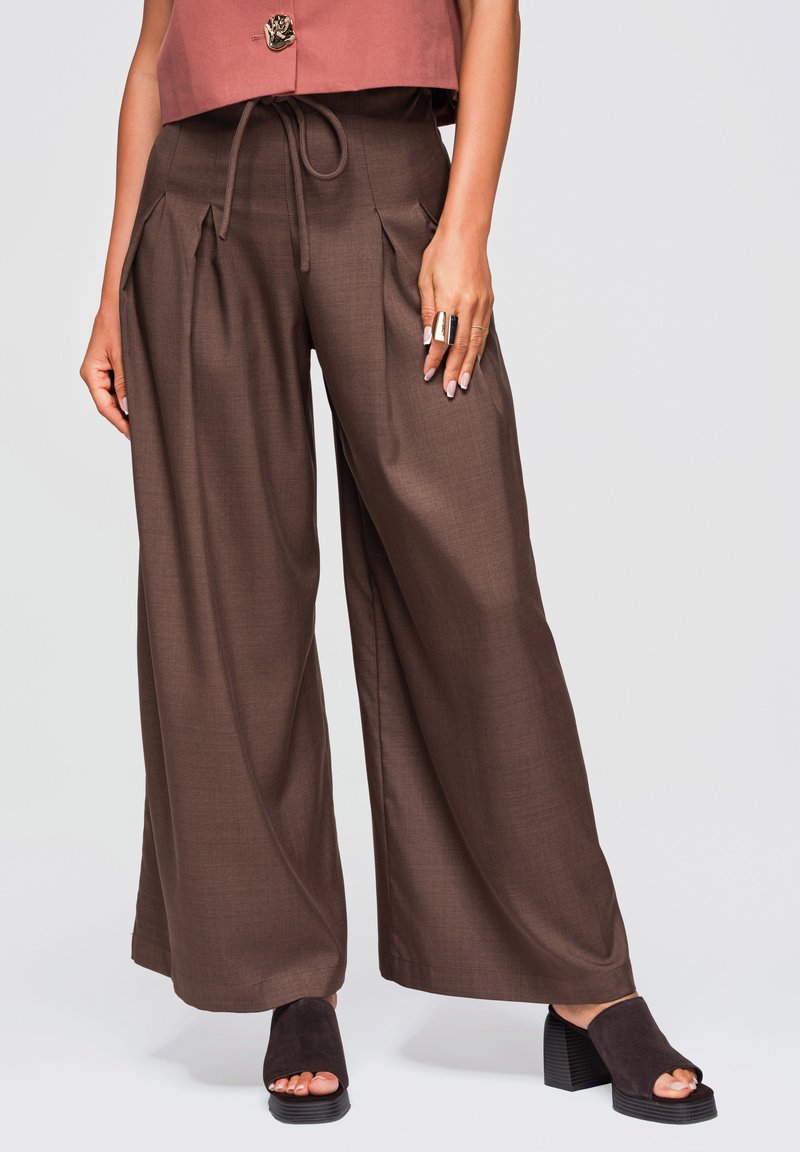 Femme portant un pantalon marron taille haute à jambes larges, avec une taille à cordon et des sandales noires à talons et orteils découverts.
