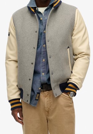 Mand iført beige og grå varsity jakke over blå denimskjorte og kaki bukser, med den ene hånd i lommen og et synligt armbåndsur.